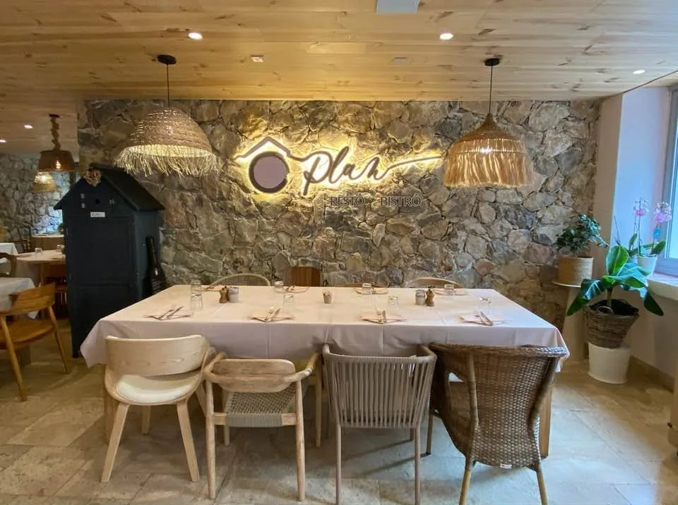 Grande table nappée devant le mur de pierre, chaises en bois et en rotin, suspensions en raphia