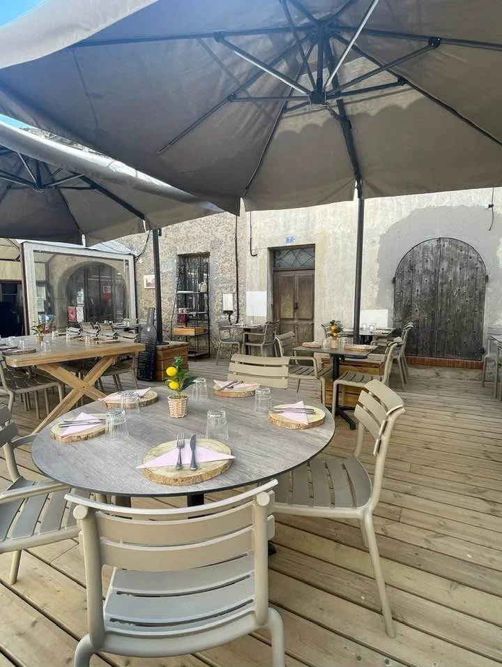 Tables rondes dressées sous de grands parasols gris, devant une porte voûtée en bois